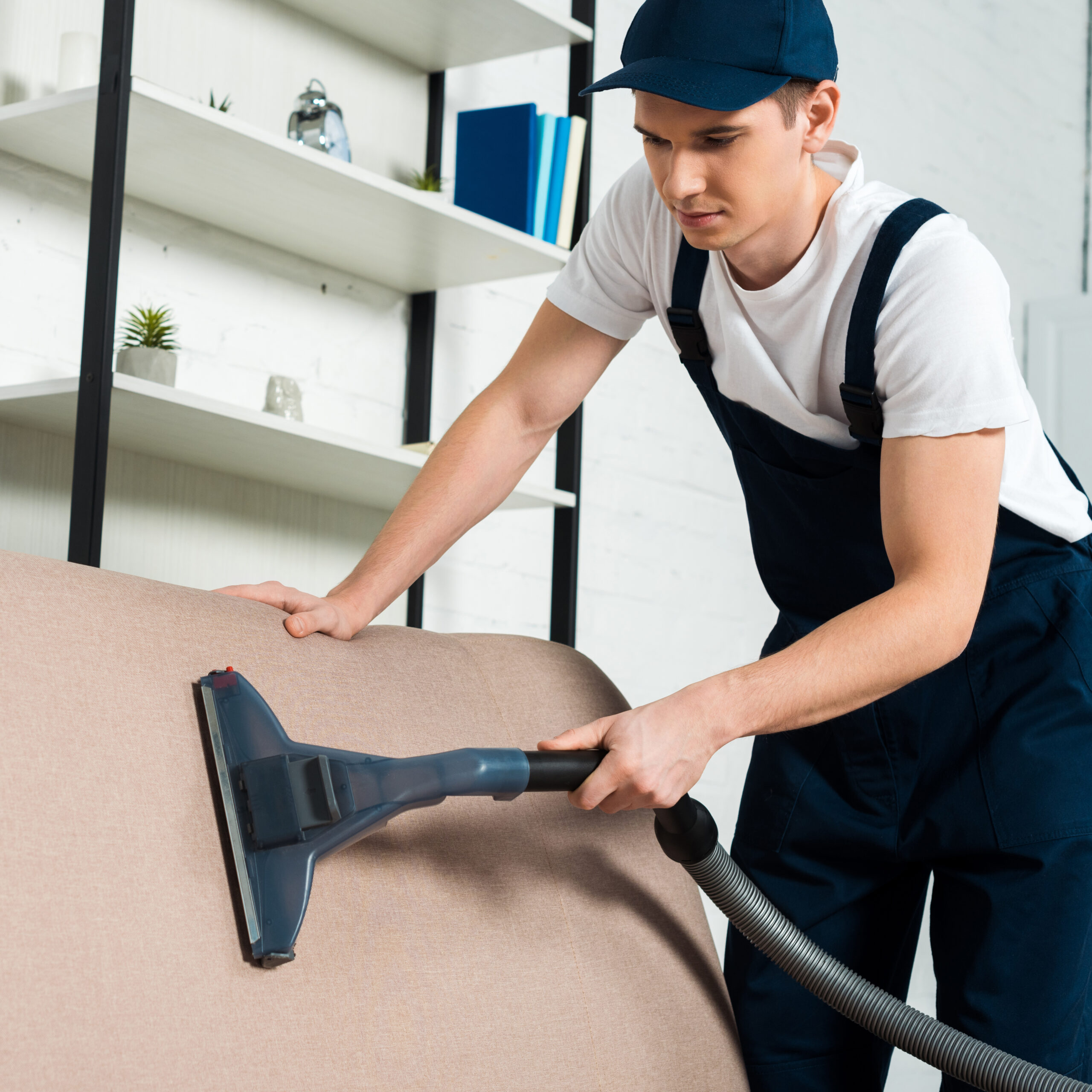 young handsome cleaner in cap removing dust on sofa with vacuum cleaner
